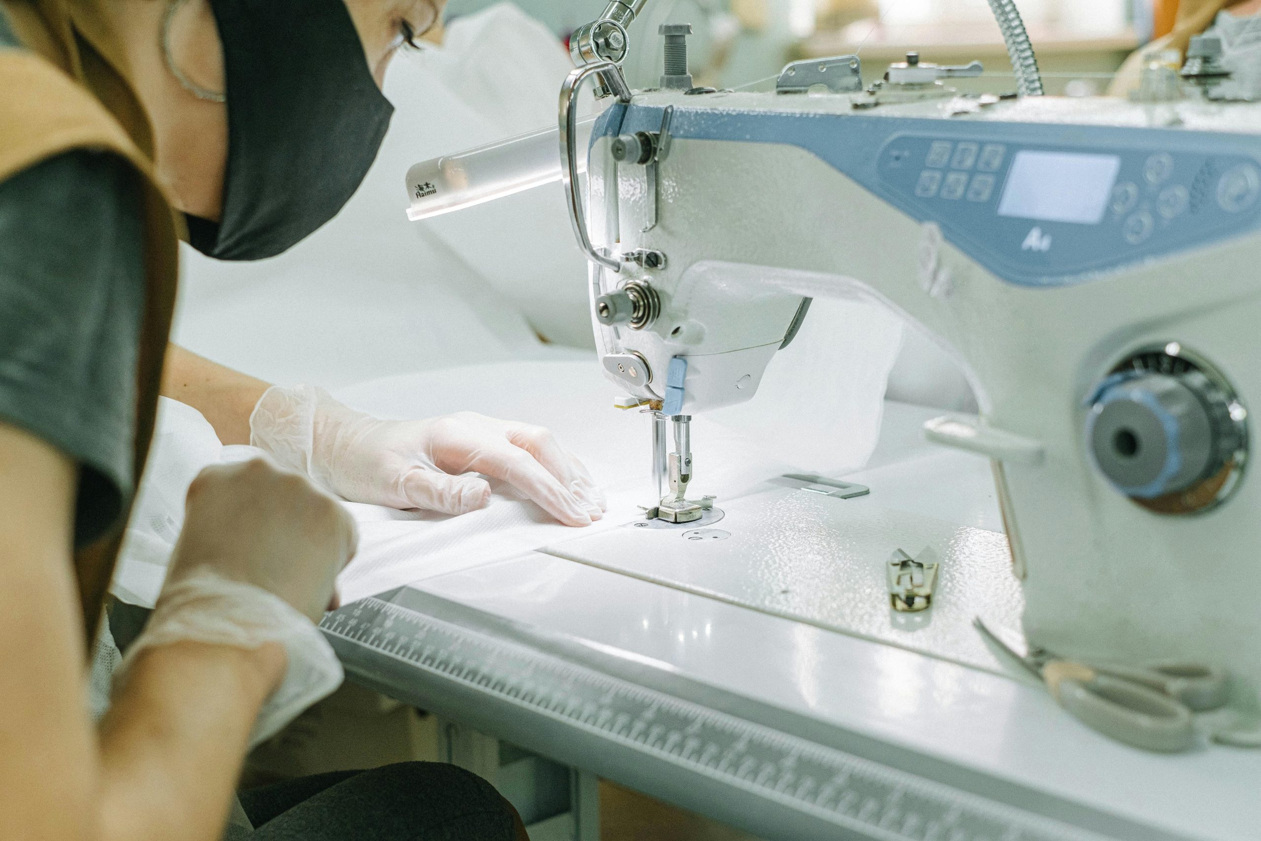 A woman wearing a face mask and gloves operates a sewing machine in a workshop.